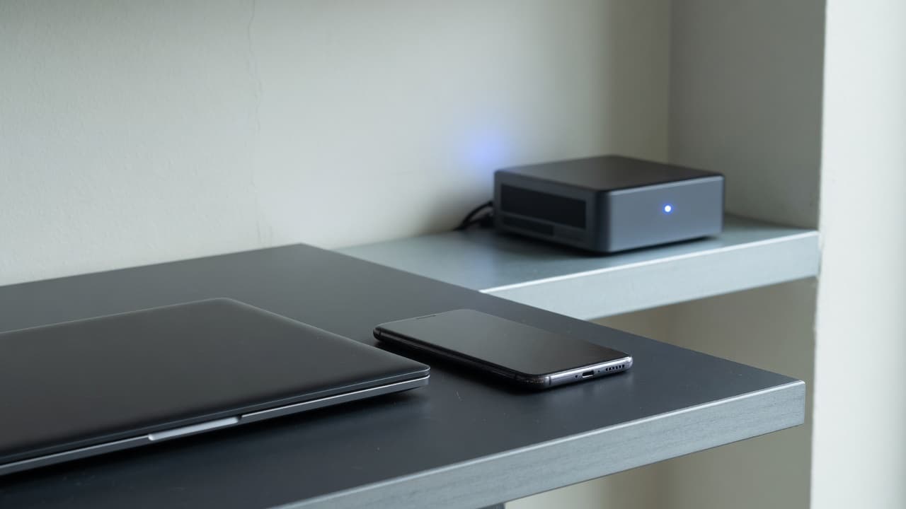 Closed laptop and phone on a desk, with a small always-on computer glowing on the shelf behind them.